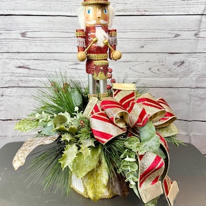 May include: A festive Christmas decoration featuring a red and gold nutcracker soldier with a white beard and a tall hat. The nutcracker stands atop a decorative arrangement of greenery, holly, and a large red and gold ribbon.