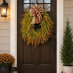 May include: A circular wreath made of green and orange foliage, featuring a large decorative bow at the top. The bow is crafted from tan burlap ribbon with a red and green plaid pattern. The wreath is hung on a dark brown wooden door with glass.