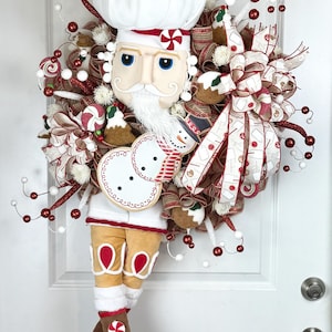 May include: A festive Christmas wreath featuring a chef design. The wreath has a white chef's hat, a face with blue eyes, and a white beard. It is decorated with red and white ribbons, candy canes, and gingerbread men. The wreath is hanging on a white door.