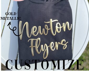 以下が含まれることがあります： 黒のフード付きスウェットシャツに、金色のメタリックな文字で「Newton Flyers」と書いてあります。テキストはカスタムエンボス加工されており、スウェットシャツはカスタマイズできます。