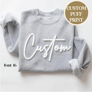 Könnte beinhalten: Ein graues Sweatshirt mit dem Wort "Custom" in weißer, gepuffter Schrift. Der Text "font 16" befindet sich unten links. Eine runde Grafik in der oberen rechten Ecke zeigt "CUSTOM PUFF PRINT".