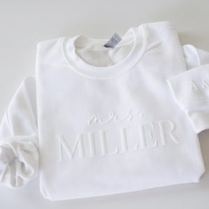 May include: White crew neck sweatshirt with the embossed text "mrs. MILLER" on the front. The sleeve cuff features the initials "A & P". The sweatshirt is folded neatly, showcasing its design.