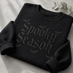 May include: A black crewneck sweatshirt featuring the embroidered text Spooky Season in a gothic font. The design includes small embroidered bat silhouettes scattered around the lettering. The garment has long sleeves with ribbed cuffs.