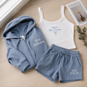 May include: A dusty blue zip-up hoodie with Mrs. Johnson embroidered on the chest, matching dusty blue shorts with EST. 8.22.25 embroidered on the leg, and a white ribbed tank top with future Mrs. Crooks and a small bow embroidered in blue.