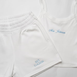 May include: White athletic shorts and a matching tank top. The shorts have an elastic waistband and the text "EST. 2.14.2026" embroidered in light blue. The tank top is embroidered with "Mrs. Johnson" in light blue.