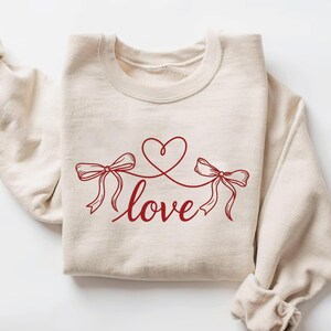 Könnte beinhalten: Cremefarbenes Sweatshirt mit roter Grafik. Die Grafik zeigt zwei Schleifen, die durch eine Linie verbunden sind, die ein Herz bildet, mit dem Wort "love" in Schreibschrift darunter. Die Ärmel sind gefaltet.