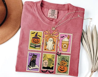 May include: A dusty rose Comfort Colors t-shirt featuring a Halloween-themed stamp design. The stamps depict a pumpkin, ghost, coffee, black cat, zombie hand, and stacked pumpkins. A silver necklace with a pendant is draped on the shirt.