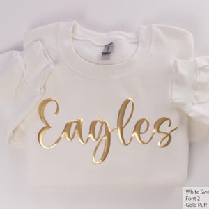 May include: White sweatshirt with the word "Eagles" written in gold puff lettering.