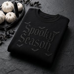 Peut inclure: Un sweat-shirt noir à col rond avec le texte Spooky Season brodé en fil noir assorti. Le motif comprend de petites silhouettes de chauves-souris entourant le lettrage, créant un aspect ton sur ton et texturé sur le tissu.