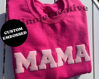 May include: Pink t-shirt with the word "MAMA" in raised pink letters. The shirt is folded and has a black circle with the text "CUSTOM EMBOSSED" on it.
