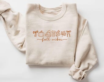 Könnte beinhalten: Ein cremefarbenes Sweatshirt mit dem Schriftzug "fall vibes" in brauner Schrift. Das Design umfasst braune Illustrationen einer Schleife, eines Kürbisses, einer Mütze, einer Kaffeetasse, eines Schals und eines Blattes. Die Ärmel sind hochgekrempelt.