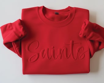 Puede incluir: Una sudadera roja brillante con la palabra "Saints" en relieve, en cursiva, en la parte delantera. Las mangas están dobladas y el cuello redondo es visible. La tela parece suave y cómoda.
