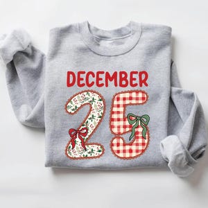 Op de afbeelding: Een lichtgrijs sweatshirt met de tekst "DECEMBER 25" in het rood. De cijfers zijn versierd met een bloemenpatroon en rode en groene strikken. Het cijfer 5 heeft een rood-wit geblokt patroon.