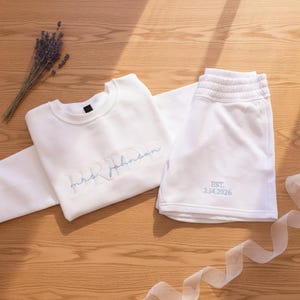 May include: A white sweatshirt and shorts set. The sweatshirt has "BRIDE" and "mrs. johnson" embroidered in light blue. The shorts are embroidered with "EST. 2.14.2026" in light blue. A sprig of lavender and a white ribbon are also visible.