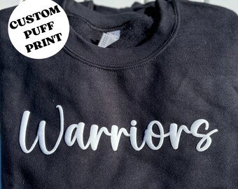 May include: Black sweatshirt with a white custom puff print that says "Warriors". The sweatshirt has a round white label that says "Custom Puff Print".