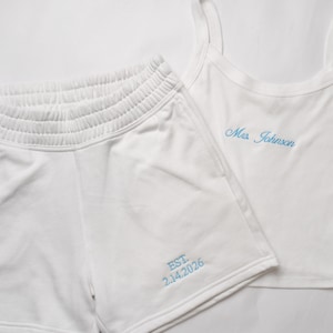 May include: White matching tank top and shorts set. The tank top has thin straps and the name "Mrs. Johnson" embroidered in light blue. The shorts have an elastic waistband and the text "EST. 2.14.2026" embroidered in light blue.