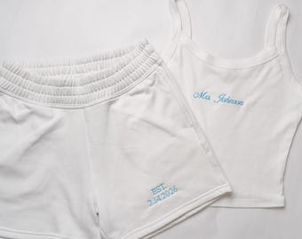 May include: White matching tank top and shorts set. The tank top has thin straps and the name "Mrs. Johnson" embroidered in light blue. The shorts have an elastic waistband and the text "EST. 2.14.2026" embroidered in light blue.