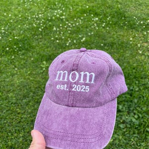 May include: Purple baseball cap with the text "mom est. 2025" embroidered in white.
