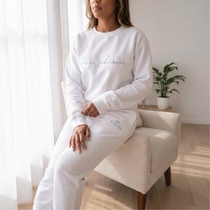 May include: White matching sweatshirt and sweatpants set. The sweatshirt has the embroidered text "Mrs. Johnson" in a light blue script. The sweatpants have the embroidered text "Est. 2.14.2026" in a light blue script.