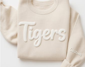 Pode incluir: Um moletom creme com a palavra "Tigers" em letras brancas em relevo no peito. O punho da manga tem a palavra "Custom" bordada em branco. O moletom está cuidadosamente dobrado.