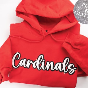 May include: A red hooded sweatshirt with the word "Cardinals" in white script with a black glitter outline. The sweatshirt has a drawstring hood and ribbed cuffs. A circular logo with the text "PUFF & GLITTER" is in the upper right corner.