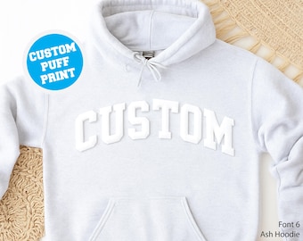 May include: Light gray hoodie with the word "CUSTOM" in white, puffy, block letters across the chest. The hoodie has a drawstring hood and a front pocket. A blue circle with the text "CUSTOM PUFF PRINT" is in the upper left corner.