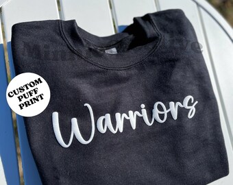 May include: Black sweatshirt with white puff print lettering that says "Warriors". The sweatshirt has a round white label that says "Custom Puff Print".