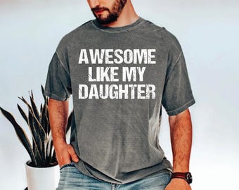 May include: A gray t-shirt with the text "AWESOME LIKE MY DAUGHTER" in white block letters. The shirt is worn by a person, and the sleeves are rolled up. The person is wearing a watch on their wrist.
