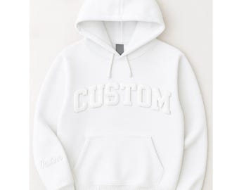以下が含まれることがあります： 白いパーカー。フードにはドローコード、カンガルーポケットが付いています。「CUSTOM」の文字が胸に浮き上がって表示されています。「Custom」の文字も左袖にあります。