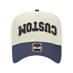 Op de afbeelding: Een witte en blauwe baseballcap met een zwart geborduurd "CUSTOM" logo op de voorkant. De cap heeft een klassieke pasvorm en een mid-profiel.