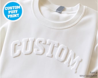 Puede incluir: Sudadera blanca con la palabra "CUSTOM" en una fuente en relieve y abultada en el pecho. Un círculo azul en la esquina superior izquierda dice "CUSTOM PUFF PRINT". El texto "Font 6 Whit Sweatshirt" está en la esquina inferior derecha.