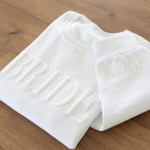 May include: White sweatshirt with the word "BRIDE" embossed across the front. The sleeve cuff of the sweatshirt features the initials "A & H". The sweatshirt is folded on a wooden surface.