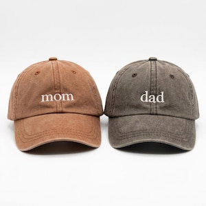 May include: Two baseball caps, one brown and one gray, are displayed. The brown cap has the word "mom" embroidered in white, while the gray cap has "dad" embroidered in white. Both caps have a curved brim and a distressed look.