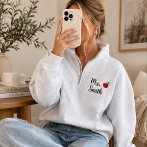 May include: A light gray quarter-zip sweatshirt featuring the embroidered black text "Ms. Smith" and a small red apple icon on the left chest. The garment has long sleeves, a high collar with a silver zipper, and a soft, heathered fabric texture.