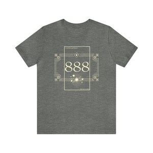 May include: A heather gray t-shirt featuring a cream-colored art deco design. The design includes the number 888, geometric shapes, and the words "Celestial guidance" and "with here as now".
