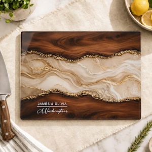 May include: A rectangular glass cutting board featuring a dark brown wood grain pattern, a central cream-coloured marble swirl design, and gold-coloured accents. The text "JAMES & OLIVIA" and "Washingtons" is printed in white on the lower left side.