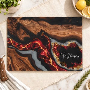 May include: A rectangular cutting board featuring a swirling pattern of dark brown wood grain, black resin, and vibrant red and gold metallic accents. The board is personalised with the white script text "The Johnsons" printed on the right side.