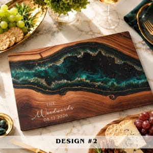A rectangular wooden serving board featuring a central inlay of dark teal and black resin with gold flecks. The board is engraved with the text "THE Woodwards 08.12.2026" in white lettering. The wood grain is visible on the left and right sides.