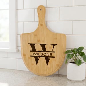 May include: A bamboo cutting board with a handle, featuring a shield shape and the engraved text "Wilsons". The board is light brown and has a hole in the handle for hanging. A small potted plant is in the background.