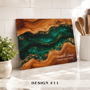 A rectangular wooden serving board featuring a central river-like inlay of swirling emerald green resin with embedded gold leaf flakes. The text "Emily & James" and "Established 2026" is engraved in white on the lower right side.