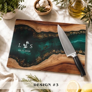 May include: A rectangular wooden cutting board featuring a central river-like inlay of dark teal resin with gold flecks. The board is engraved with the letters L and S, a small leaf icon, and the date 08.12.2026. A stainless steel chef knife rests on top.