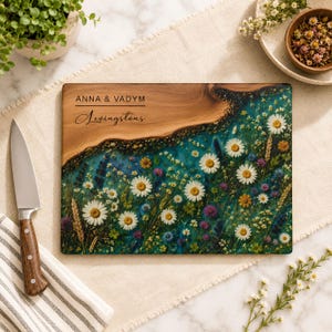 May include: A rectangular wooden cutting board featuring a natural live edge and a resin inlay filled with pressed wildflowers in shades of white, blue, and purple. The board is personalized with the text ANNA & VADYM and Levingstons.