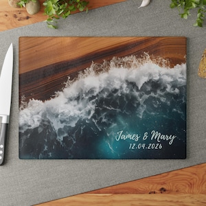 May include: A rectangular wooden cutting board featuring a printed design of ocean waves crashing onto a sandy shore. The text James & Mary and the date 12.04.2026 are printed in white script on the lower right side of the board.