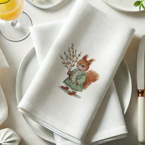 May include: White linen napkin with an embroidered design of a squirrel holding a sprig of willow branches. The squirrel is wearing a green jacket and has a brown tail.