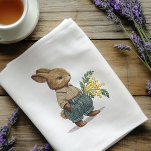 May include: A white tea towel with an embroidered design of a brown rabbit holding a bouquet of yellow mimosa flowers.