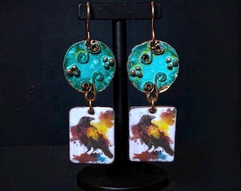 May include: Dangle earrings featuring a circular turquoise-colored top piece with embossed swirl patterns and a rectangular white bottom pendant displaying a colorful painted raven illustration with yellow, orange, and brown accents.