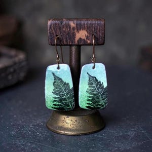 Puede incluir: Un par de pendientes rectangulares colgantes con un degradado de verde claro a verde oscuro y un estampado de silueta de hoja de helecho negra. Los pendientes cuelgan de ganchos metálicos finos en un soporte de madera en forma de T.