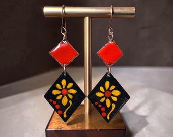 May include: A pair of dangle earrings featuring two stacked diamond-shaped charms. The top charm is solid red, while the bottom charm is black with a yellow and red flower design and three red dots. The earrings hang from gold-toned metal hooks.