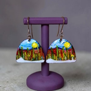 May include: A pair of dangle earrings featuring a semi-circular design with a hand-painted landscape scene. The artwork depicts a bright yellow sun in a blue sky above red and green vertical shapes, finished with a white base and metal hooks.