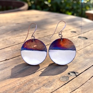 Może przedstawiać: Para ręcznie robionych kolczyków z miedzi. Każdy kolczyk ma półokrągły kształt, z białą dolną połową i górną połową, która przechodzi od brązu do niebieskiego. Kolczyki mają miedziane haczyki i są prezentowane na drewnianej powierzchni.